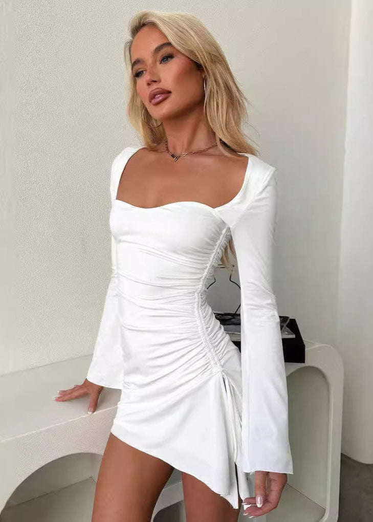 Woman with blonde wavy hair wearing a white ruched mini dress with flared sleeves and a sweetheart neckline, leaning on a white surface.