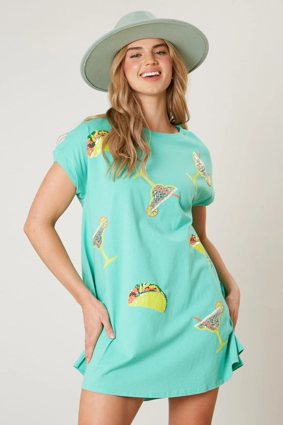 A woman wearing a mint green t-shirt dress with sequined taco and margarita prints and a light green hat, smiling.