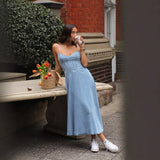 woman in a blue denim midi dress with spaghetti straps and a sweetheart neckline, holding a coffee cup and a straw tote bag filled with orange tulips, leaning against a stone bench outdoors