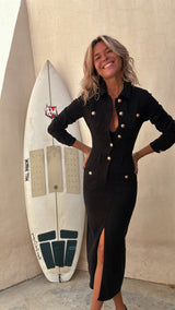 Woman in a black denim dress and gold heels standing next to a surfboard