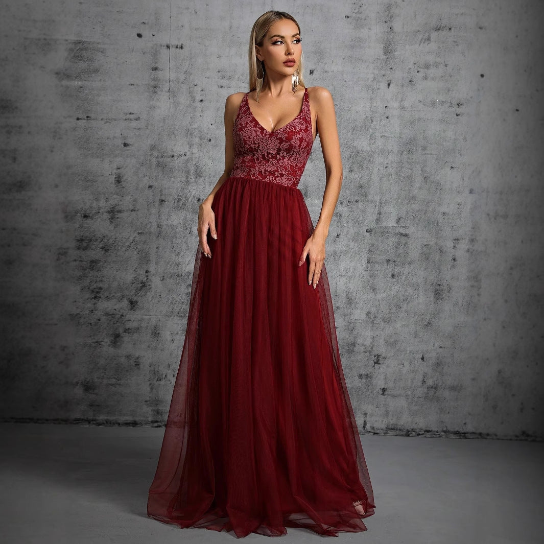 A woman in a long, flowing, deep red evening gown with a lace bodice and a V-neckline stands against a textured gray wall.