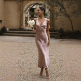 Woman in a pink satin slip dress holding a bouquet of flowers, standing outdoors with an arched walkway in the background.