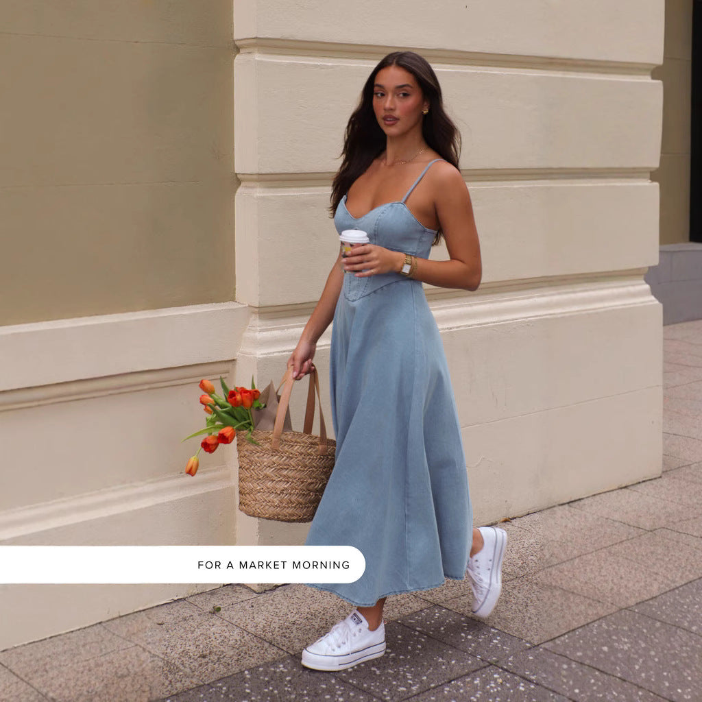 woman walking on a sidewalk wearing a blue denim midi dress with spaghetti straps and a sweetheart neckline, carrying a straw tote bag with orange tulips and a coffee cup