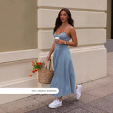 woman walking on a sidewalk wearing a blue denim midi dress with spaghetti straps and a sweetheart neckline, carrying a straw tote bag with orange tulips and a coffee cup