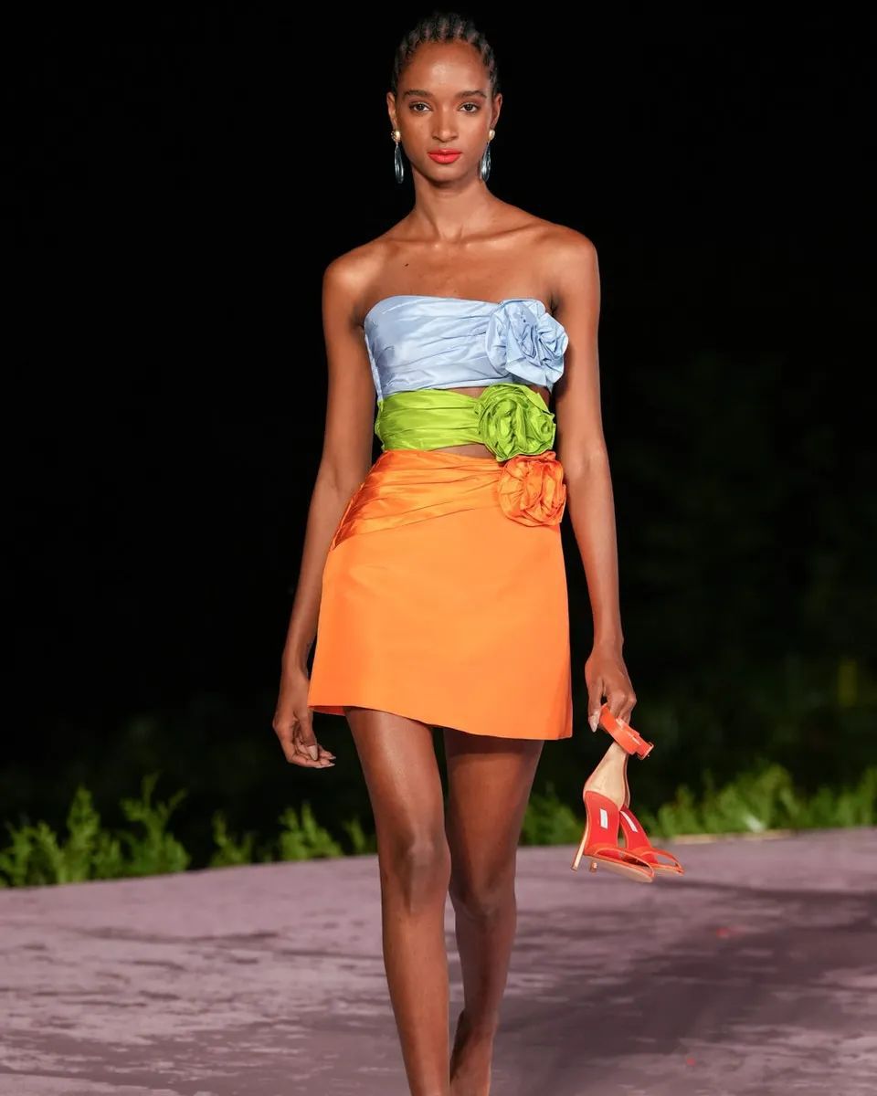 A model walks down a runway wearing a colorful strapless dress with floral embellishments.