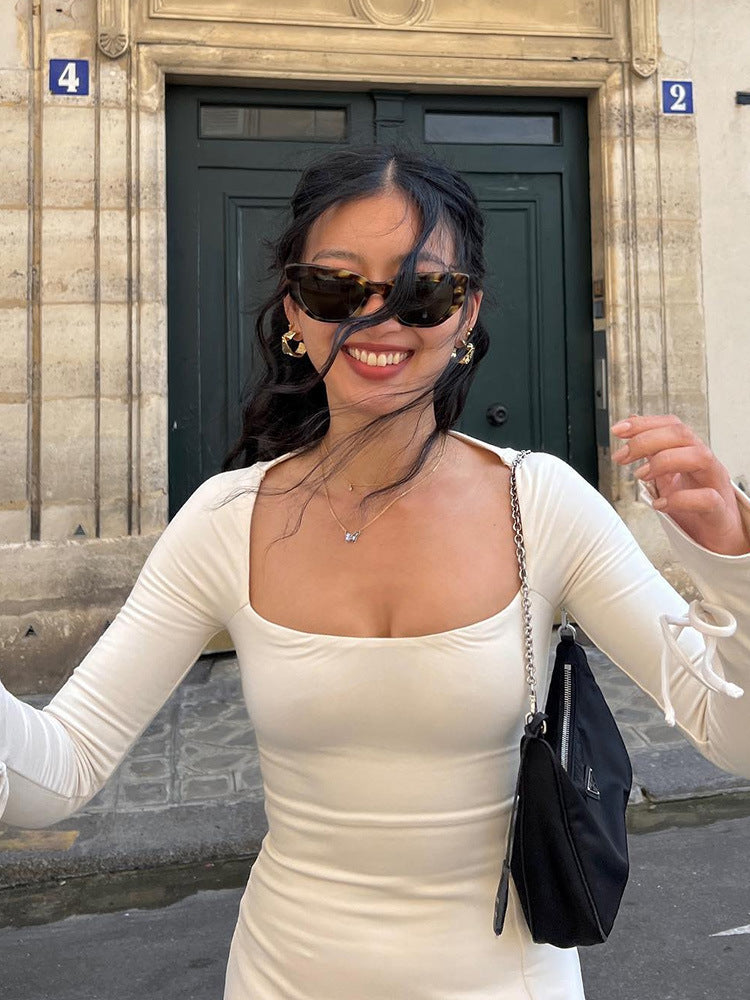 A woman in an ivory long-sleeved mini dress with a square neckline and tie details on the sleeves smiles at the camera.