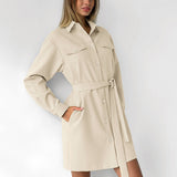 Woman wearing a long-sleeved beige shirt dress with a belted waist and chest pockets, leaning against a white wall with one hand in her pocket.