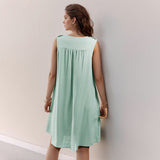 The back view of a young woman wearing a mint green sleeveless A-line dress with a button-front and pleated yoke. She is standing against a light-colored wall.