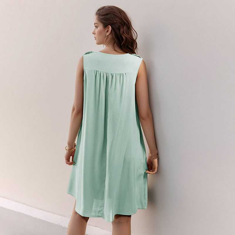 The back view of a young woman wearing a mint green sleeveless A-line dress with a button-front and pleated yoke. She is standing against a light-colored wall.
