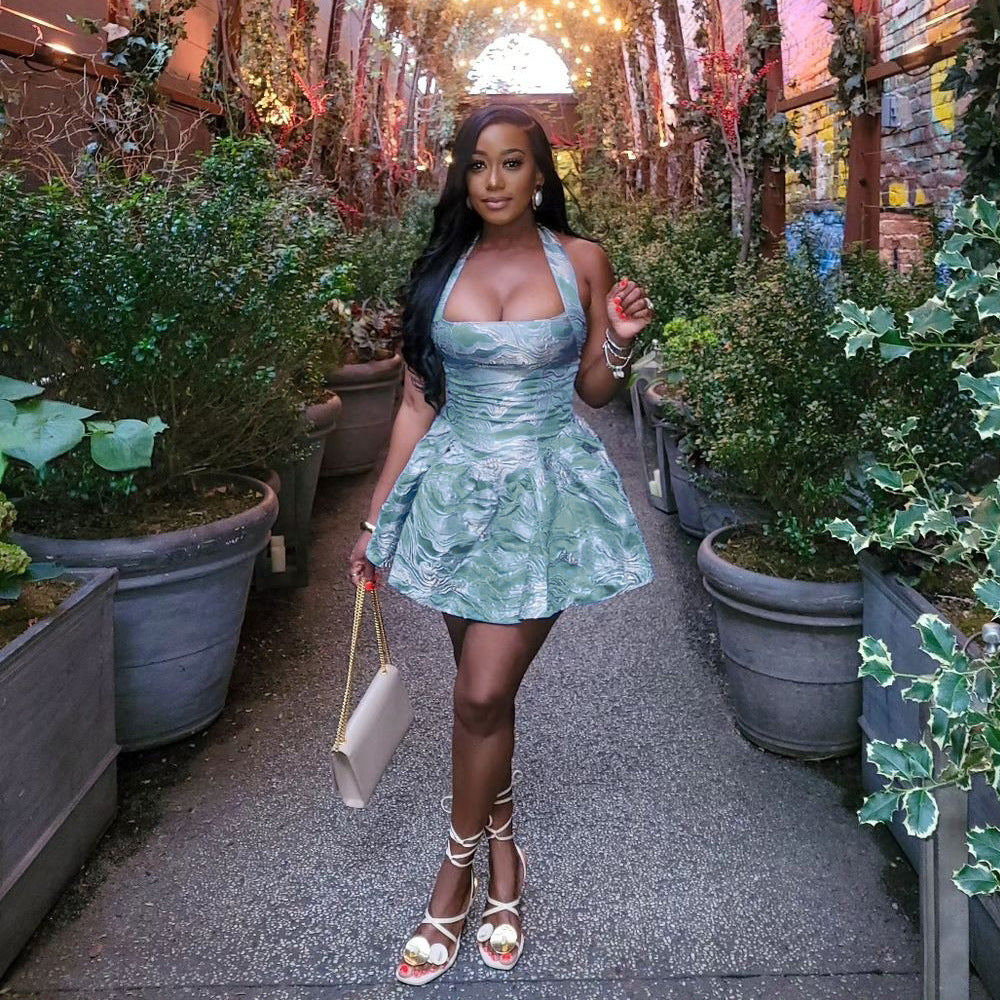 A woman in a green halter dress with a pleated skirt and white strappy heels poses in a garden.