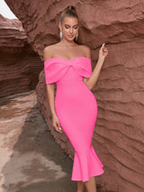 A woman in a vibrant pink off-the-shoulder dress with a large bow, looking down with her hand gently touching her chest, set against a rustic rock formation.