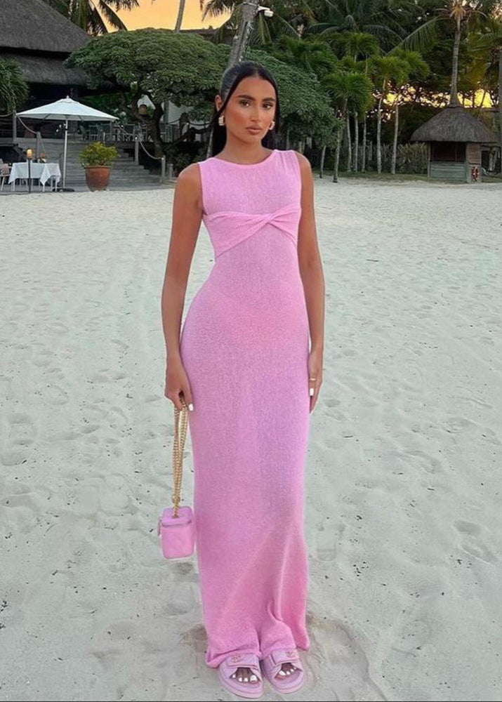 A woman in a pink knit maxi dress stands on a beach with a small pink handbag.