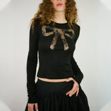 A woman wears a black long-sleeved shirt with a lace bow design on the chest and a black ruffled skirt.