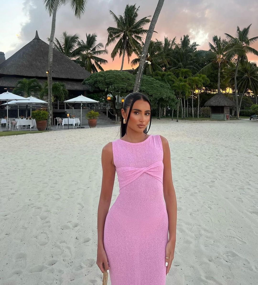 A woman in a pink maxi dress stands on a beach at sunset.