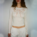 A woman wears a cream-colored long-sleeved shirt with a lace bow design on the chest and white drawstring pants.