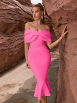 A woman wearing a bright pink off-the-shoulder dress with a bow detail and a mermaid hem, posing in a rocky, desert-like environment.