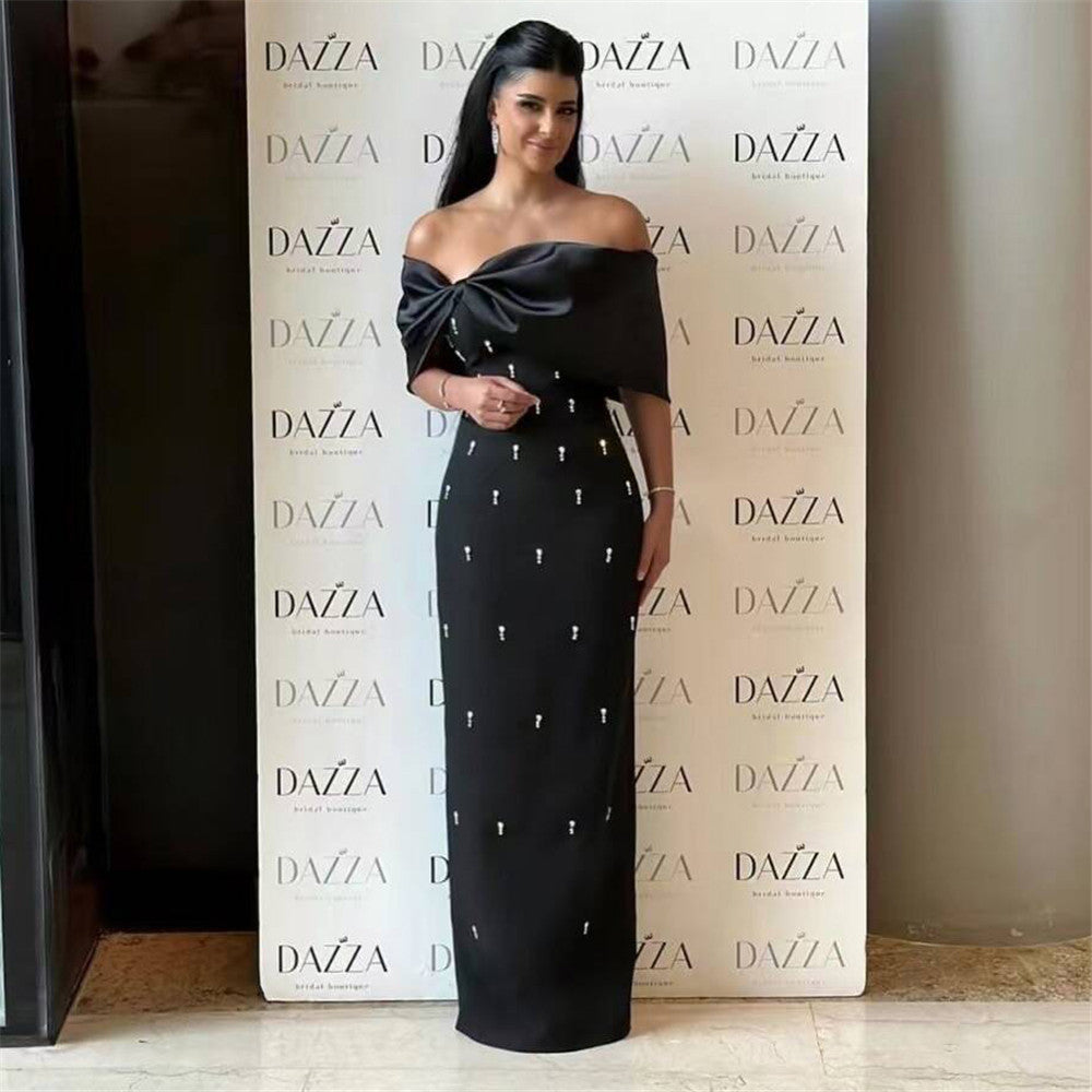 A woman in a black off-the-shoulder gown with a large bow on the bodice and small white embellishments scattered throughout the skirt, standing in front of a white backdrop with a repeated logo.