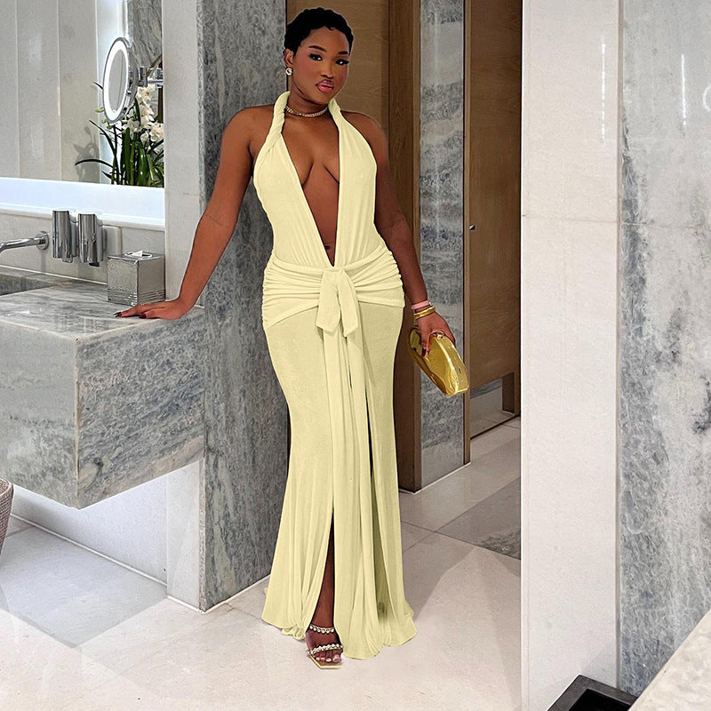 A woman in a long, light yellow halter neck dress with a deep V-neck and a high slit, leaning against a bathroom counter.