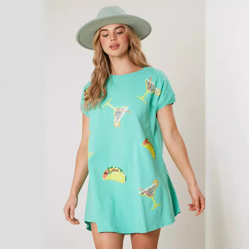 A woman wearing a mint green t-shirt dress with sequined taco and margarita prints and a light green hat.