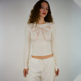 A woman wears a cream-colored long-sleeved shirt with a lace bow design on the chest and white drawstring pants.