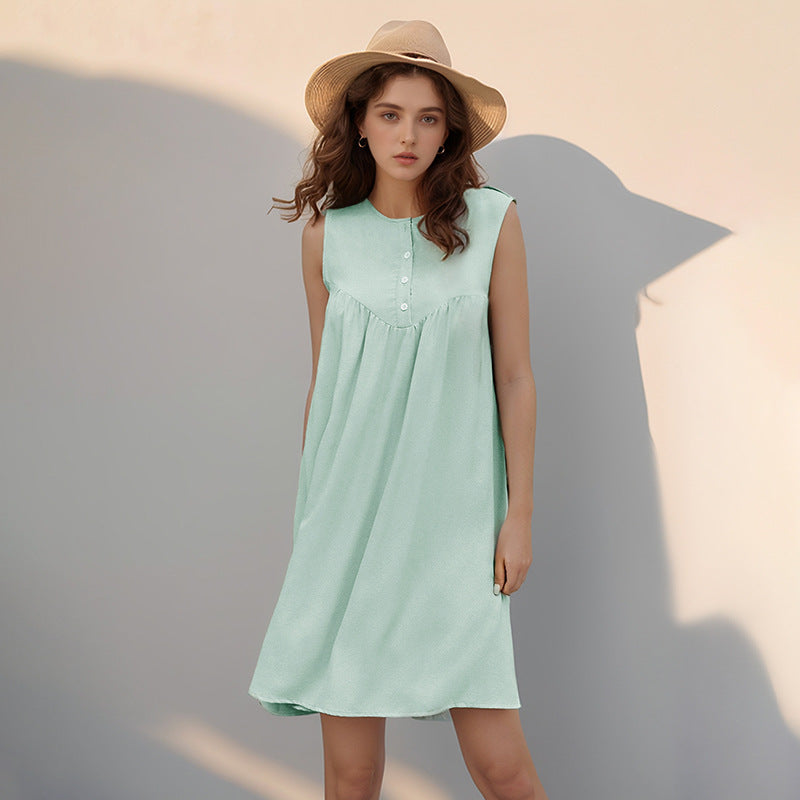 A young woman wearing a mint green sleeveless A-line dress with a button-front and pleated yoke poses against a light-colored wall. She is wearing a straw hat.