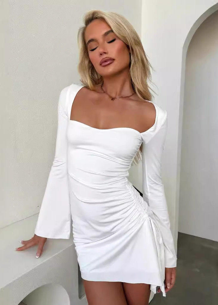 Woman leaning against a white surface, wearing a white ruched mini dress with flared sleeves and a sweetheart neckline.