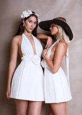 Two women wearing matching white halter neck dresses with deep V-necklines and rose embellishments at the waist. One wears a flower crown, the other a large black hat.