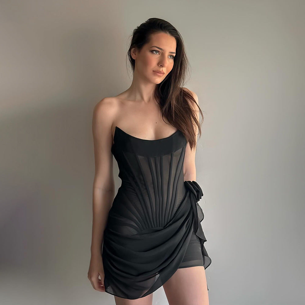 A woman in a black strapless corset dress with sheer fabric and a draped skirt, posing against a gray wall.