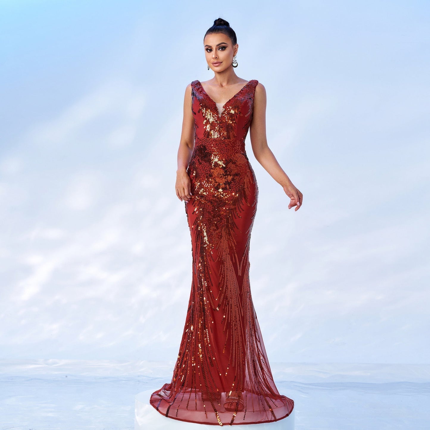 Woman in a red sequined mermaid gown with a v-neck and sleeveless design, posing on a white platform.