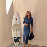 Woman in a denim dress and cowboy hat standing next to a surfboard