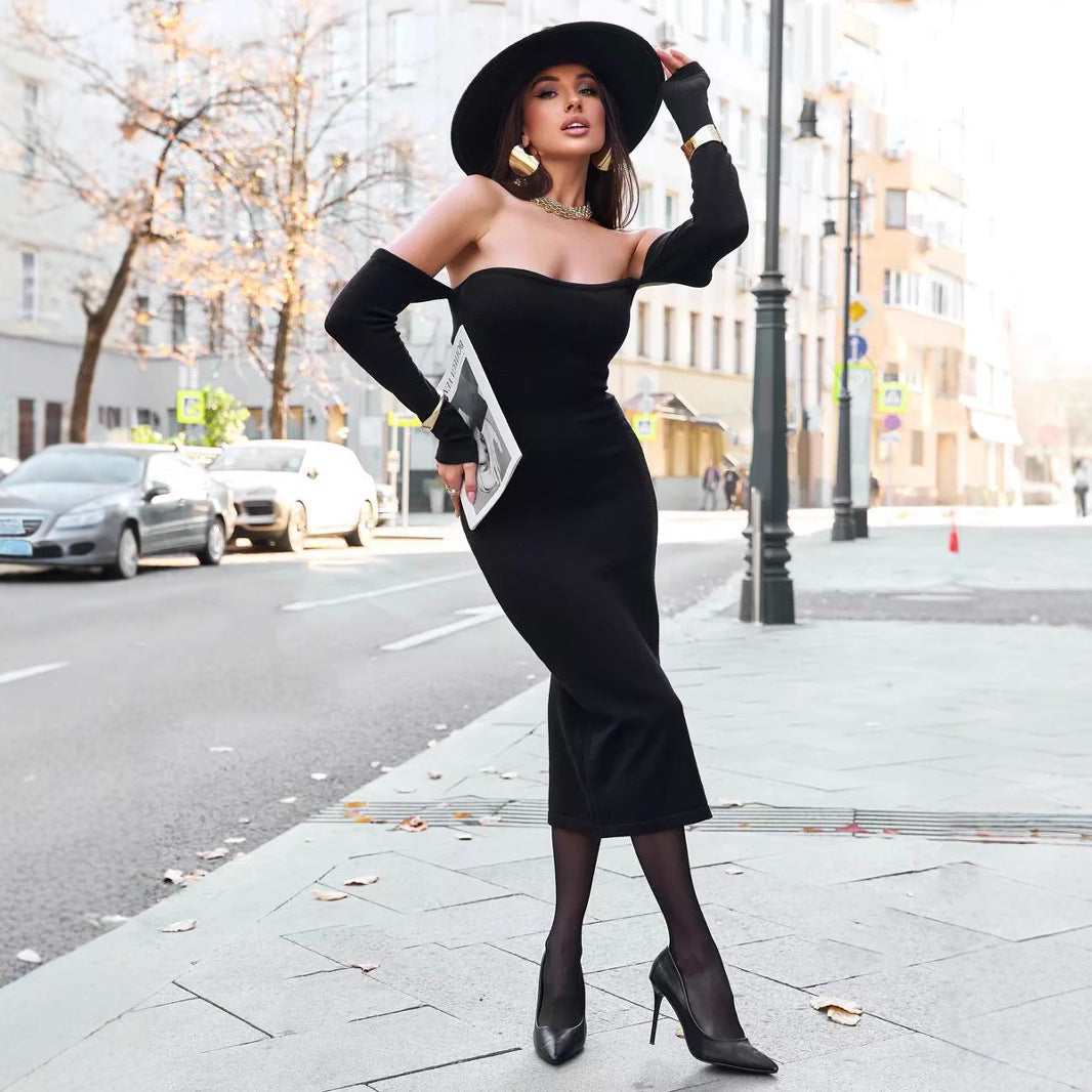 A woman in a black off-the-shoulder midi dress, black hat, and high heels stands on a city sidewalk.