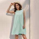 A young woman wearing a mint green sleeveless A-line dress with a button-front and pleated yoke poses against a light-colored wall. She is wearing a straw hat and adjusting it.