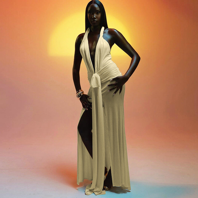 A woman in a long, beige halter neck dress with a deep V-neck and a high slit, posing against a gradient background.