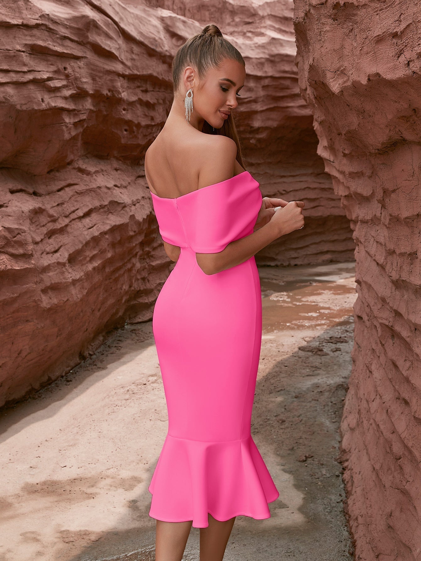 Rear view of a woman in a form-fitting hot pink off-the-shoulder dress with a mermaid flare, standing in a narrow canyon.