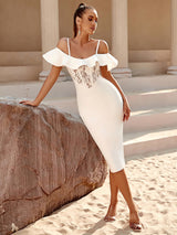 A woman in a white off-the-shoulder midi dress with a lace bust panel leans against a large rock on a sandy surface, with stone columns in the background.