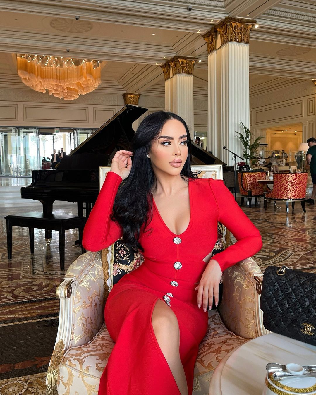 Woman with long black hair wearing a red long-sleeved midi dress with a high slit, sitting in a luxurious hotel lobby.