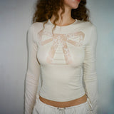 A woman wears a cream-colored long-sleeved shirt with a lace bow design on the chest and white drawstring pants.