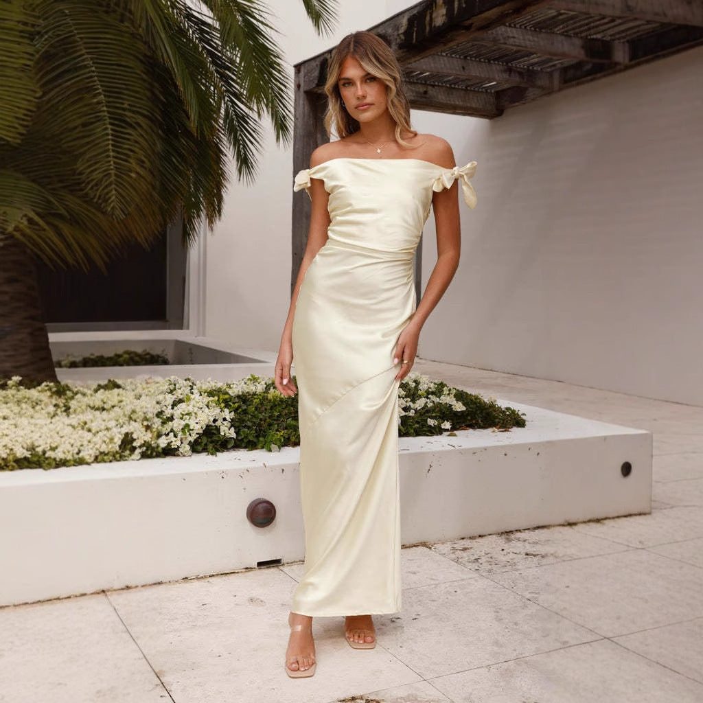 Woman in a cream off-the-shoulder satin gown with bow accents on the shoulders, standing outdoors.