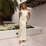Woman in a cream off-the-shoulder satin gown with bow accents on the shoulders, standing outdoors.
