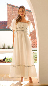 A woman models a long, white, ruffled dress with a square neckline and puff sleeves.