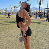 A woman in a black one-shoulder top and mini skirt poses from the side at an outdoor festival with palm trees and a Ferris wheel in the background.