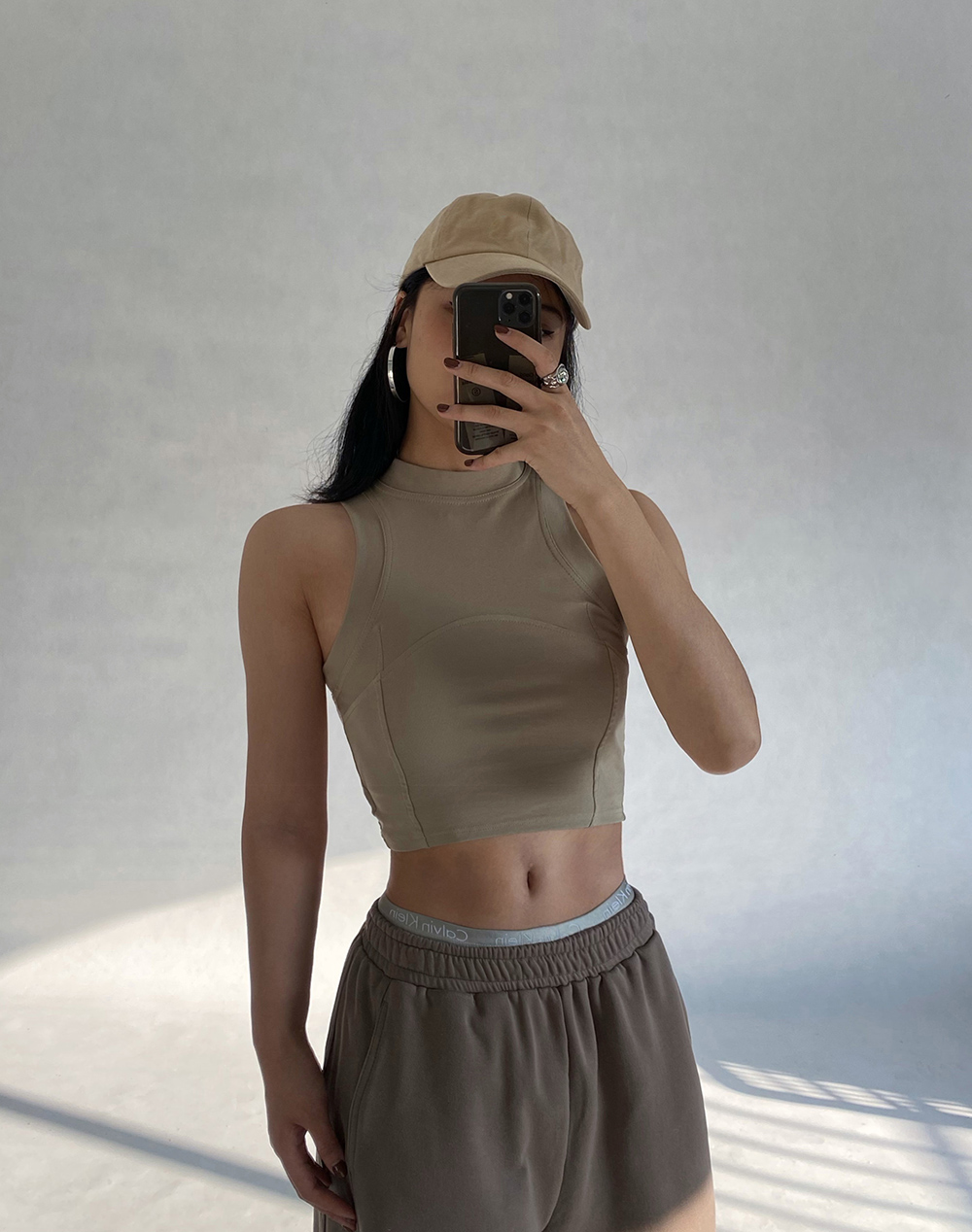 Woman wearing a beige cropped tank top, brown sweatpants, and a beige baseball cap, holding a phone to take a selfie.