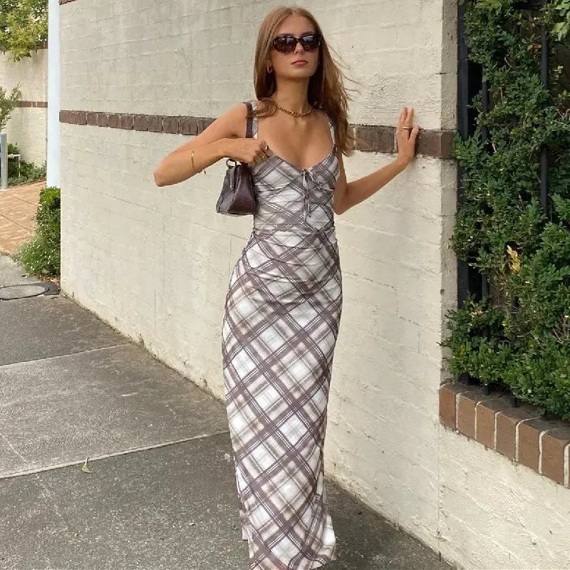 A young woman leans against a brick wall, wearing a brown and white plaid maxi dress, sunglasses, and a brown shoulder bag.