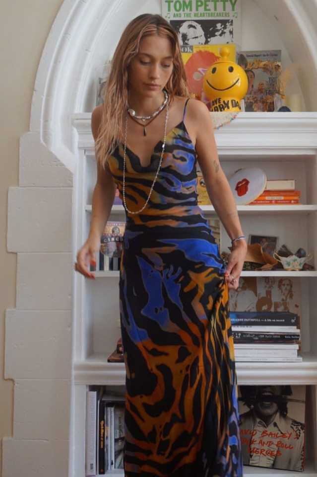 A woman in a long, flowing, animal-print dress stands indoors in front of a bookshelf.