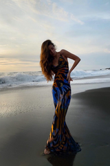 A woman in a long, flowing, animal-print dress poses on a black sand beach with waves in the background.
