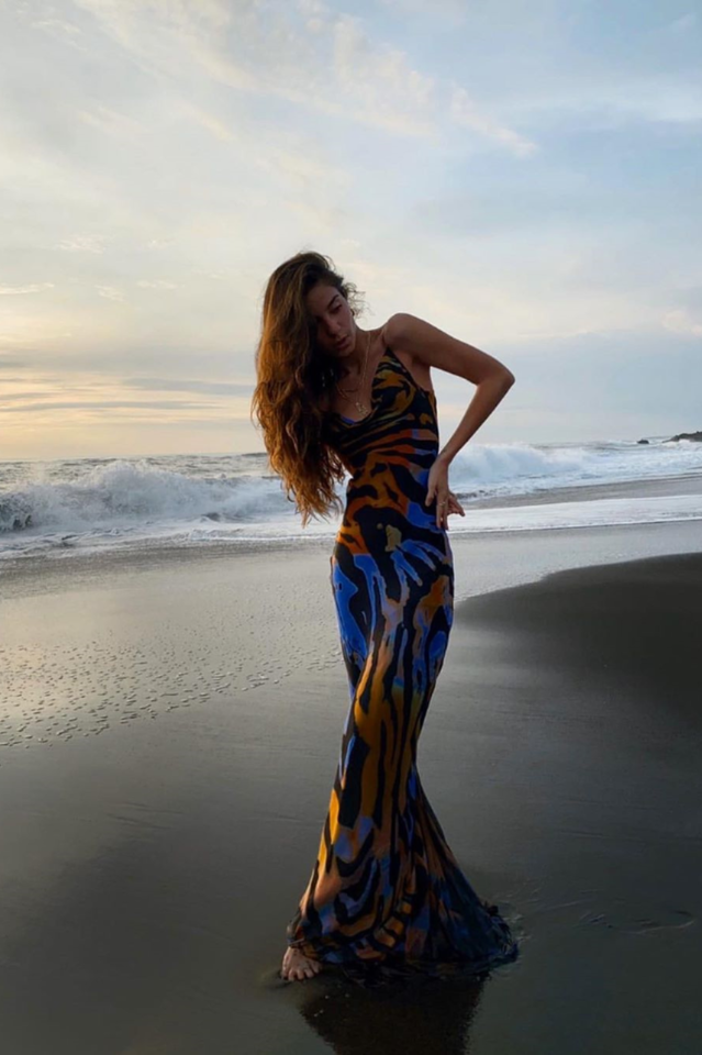 A woman in a long, flowing, animal-print dress poses on a black sand beach with waves in the background.