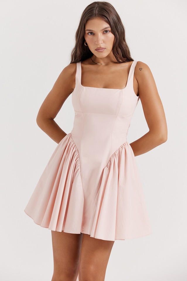 Woman in a pink mini dress with a ruched skirt and square neckline, posing with hands on hips.
