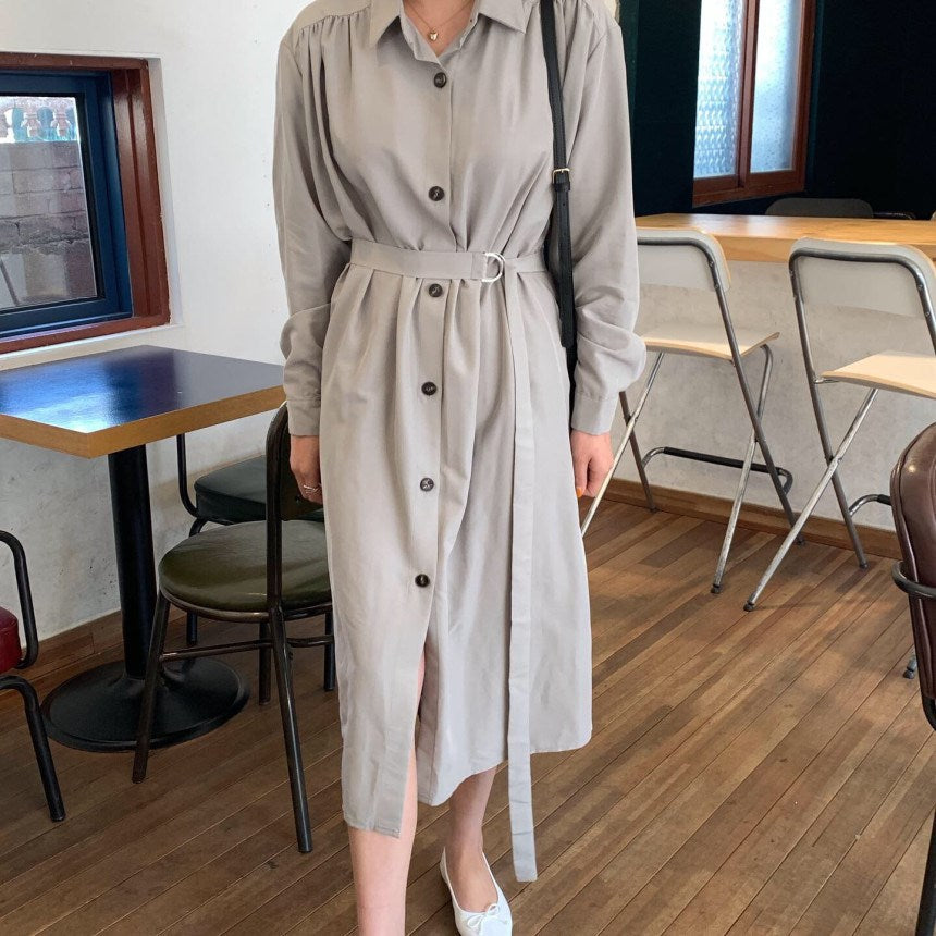 Woman wearing a long gray button-down dress with a belt and white flats, standing in a cafe.