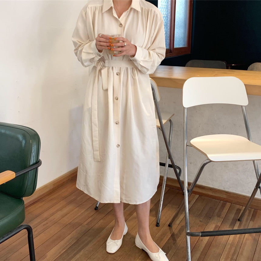 Woman in a cream-colored button-down dress and white flats, holding a drink.