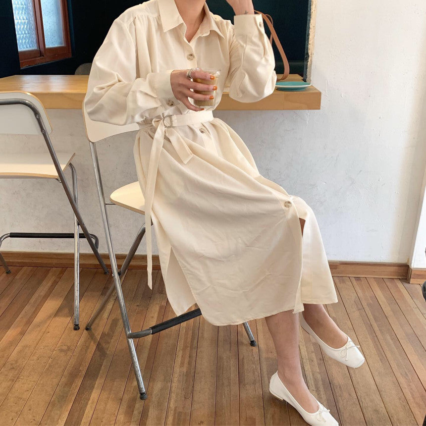 Woman sitting on a chair in a cream-colored button-down dress and white flats, holding a drink.